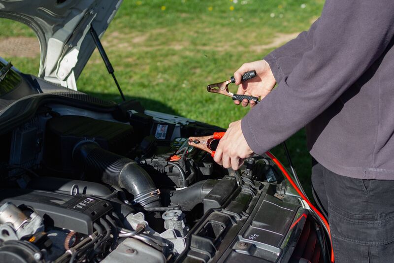 A person clamping jumper cables onto a car battery under an open hood