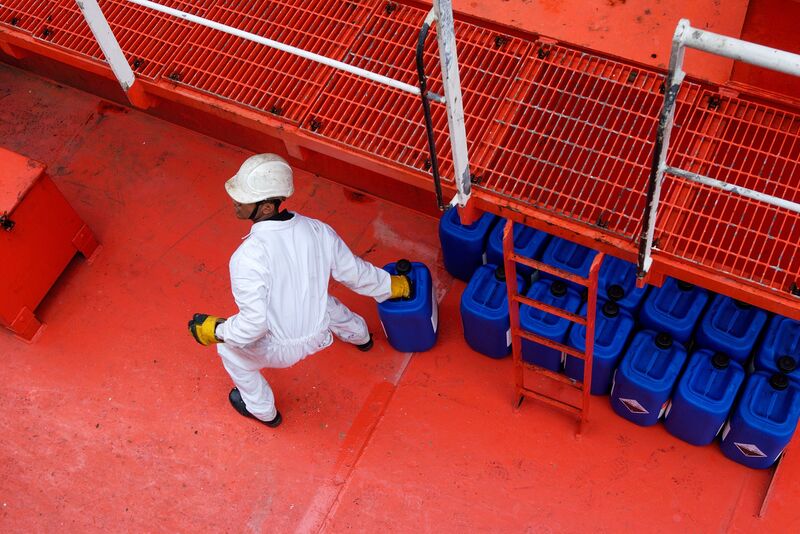A worker in coveralls carrying a jerry can past rows of stacked fuel canisters
