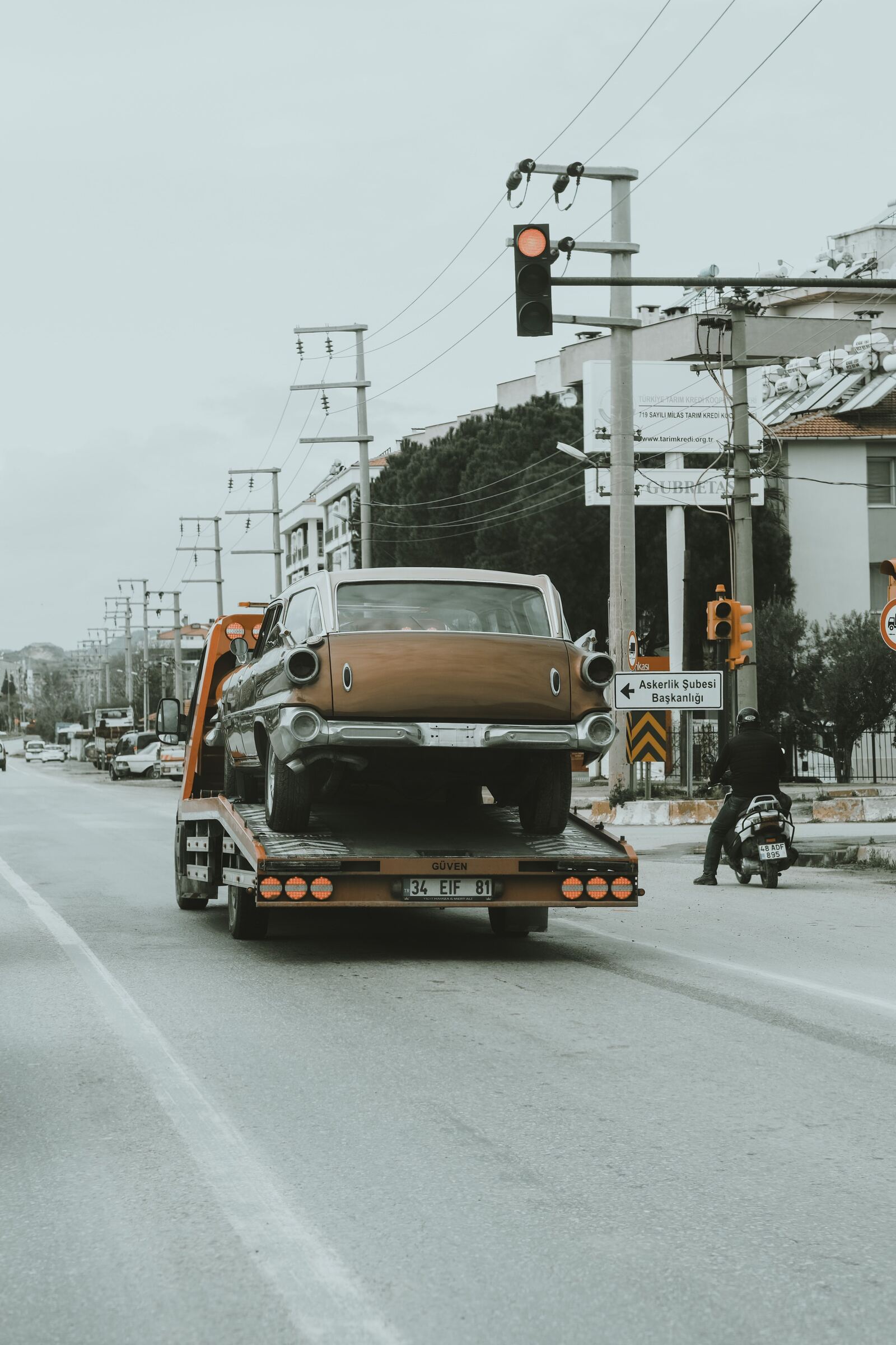 A classic car strapped to the bed of a flatbed tow truck being driven along a main road