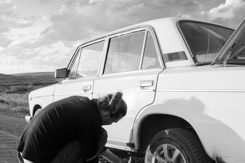 A person kneeling by a car’s rear wheel to change a flat tire at the roadside